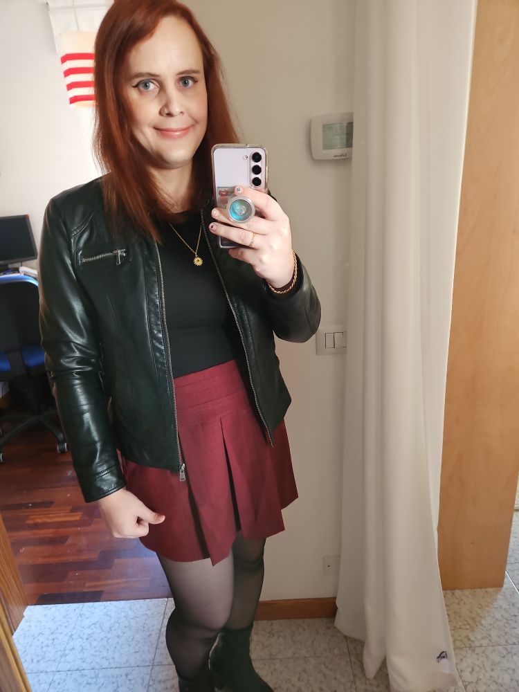 A picture of a woman taking a selfie in a mirror. She has long red hair and blue eyes and is wearing a maroon miniskirt with black tights, a black shirt, and a leather jacket on top.