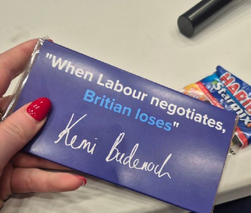A chocolate bar custom-printed for the Conservative Party conference. Text reads “When Labour negotiates, Britian loses”. That’s B R I T I A N. Wrapper is purple, text is white with the last two words in pale blue. Bar is held from leftbif frame by a hand with red nails, in front of a cream table top. 