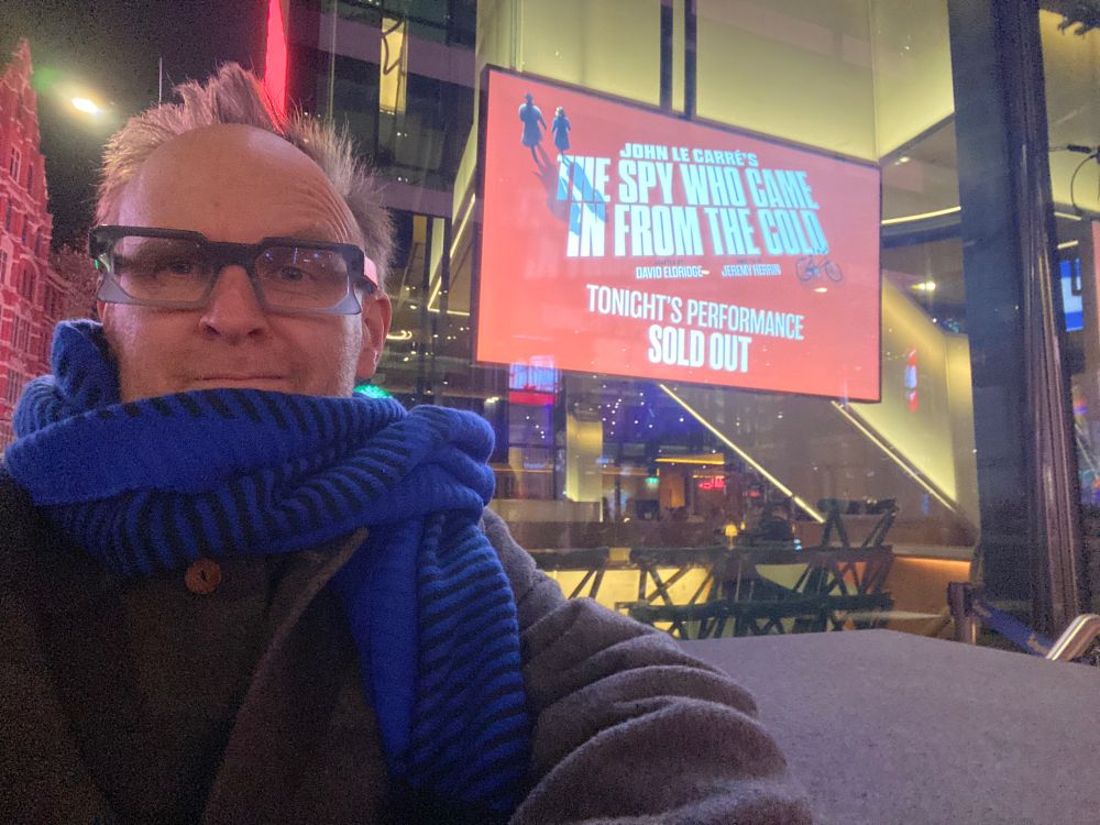 Me outside Soho Place Theatre, where the white-on-red digital poster for The Spy Who Came In From The Cold glows in the dark. It’s night and I’m wrapped up warm: grey coat, blue scarf up around my lower face, glasses and spiky dark blond hair. 
