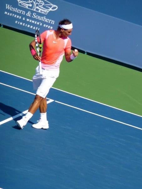 Rafa in white knee length shorts, not quite pirate pants. He has on white shoes and socks with orange shirt with some white on sides and shoulders. White bandana. He’s doing a positive fist pump while taking steps towards middle of the court. In background is the logo for The Western and Southern Open tournament which takes place in Mason, Ohio. Rafa stands out on his orange & white on the bright blue and green courts. He is giving off positive energy. Sunny day. Very bright. 