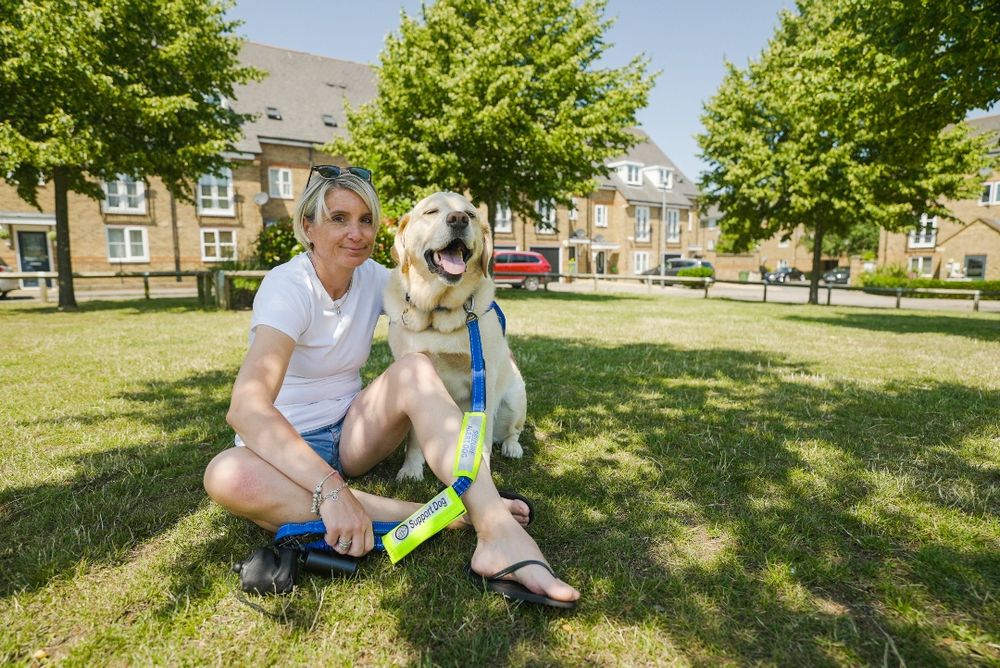A woman sits on the grass with Cobble, a yellow labrador assistance dog by her side