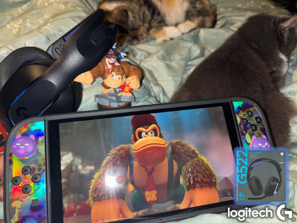 POV Saffista’s lap with a mint green comforter, two cats, a Nintendo Switch 2, a DK amiibo figure, and the LogitechG G522 headset in black. 