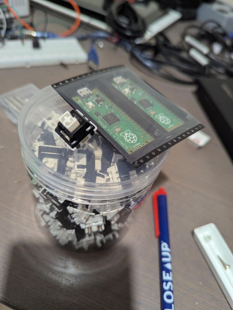 On top of a messy table is a plastic container full of keyboard switches with one switch resting on the lid, plus a double pack of Raspberry Pi Pico boards. There's a breadboard and assorted junk in the background 