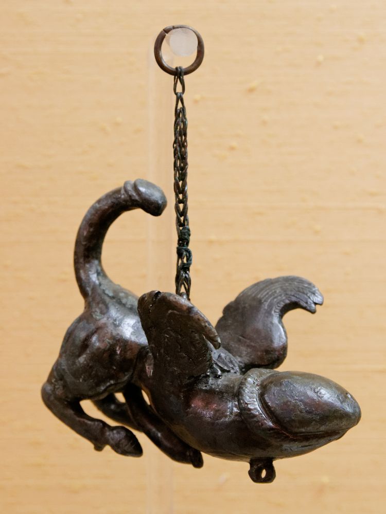 A small metal amulet of a two legged phallic figure with wings and a penis tail. It hangs from a chain.