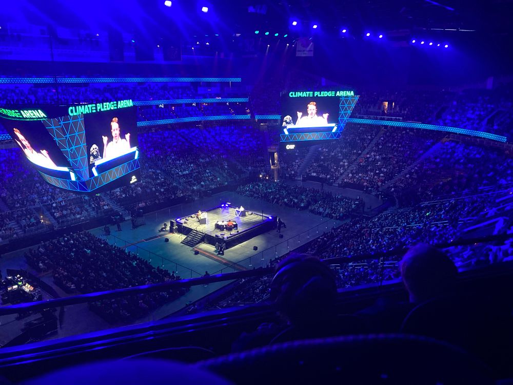 Photo from the seats at Climate Pledge Arena in Seattle of Dimension 20 Live. Brennan can be seen on the large screen with his characteristic animated gestures. The Intrepid Heroes can be seen lit in the center of the arena while a deep dark blue glow covers the audience. The energy is wild and the vibes are immaculate. 
