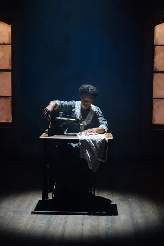 In a darkened room, Samira Wiley as Esther works at a sewing machine. Intimate Apparel at the Donmar Warehouse, 2025.