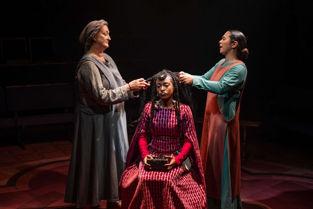 In a red Mediaeval gown, Arsema Thomas as Clare sits grumpily while Jacoba Williams and Liz Kettle as her servants braid her hair. Poor Clare at the Orange Tree Theatre, 2025.