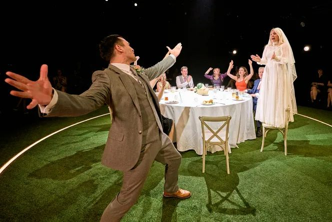 Around a circular formal table, Sinéad Matthews as Sylvia wears a wedding dress and stands on a chair, while in the foreground Julian Kostov as Marek wears a suit and stands with his arms and legs wide, looking at her. Till The Stars Come Down at the Theatre Royal Haymarket, 2025.
