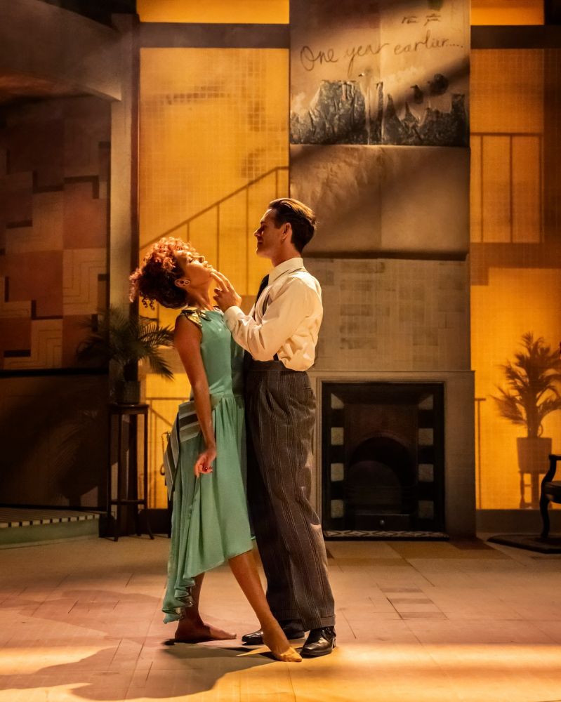 Emma McDonald as Marie-Louise and Luke Norris as John stand close to each other against a sepia-toned Art Deco background, and he touches her face. The Constant Wife at the Swan Theatre, Stratford-upon-Avon, 2025.