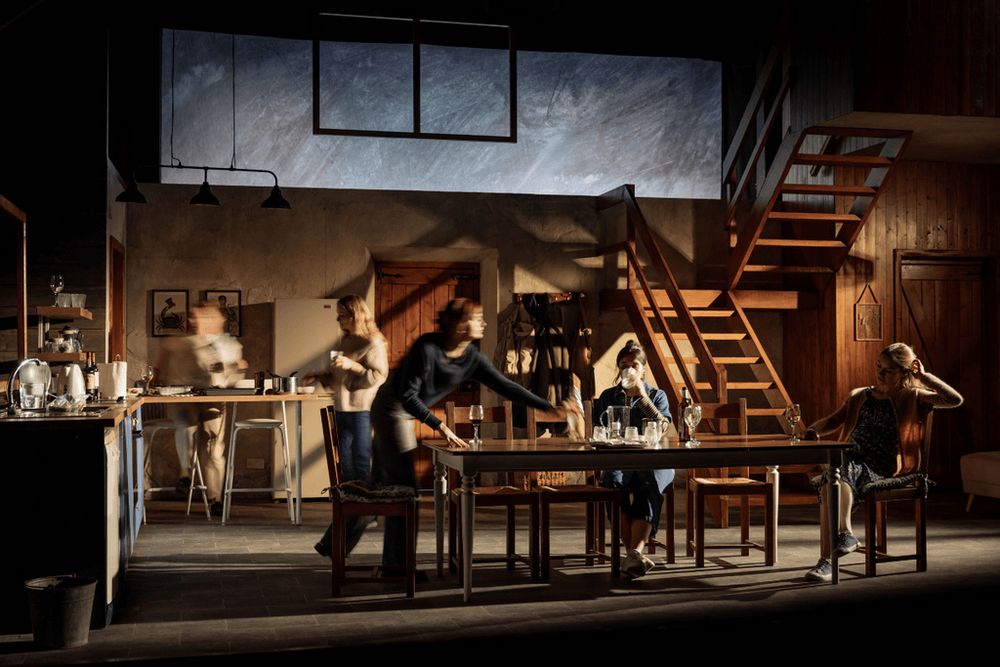 On a dimly lit set of a kitchen with wooden stairs leading up, the cast of Reunion are blurry as they move around the stage. Reunion at the Kiln Theatre, 2025.