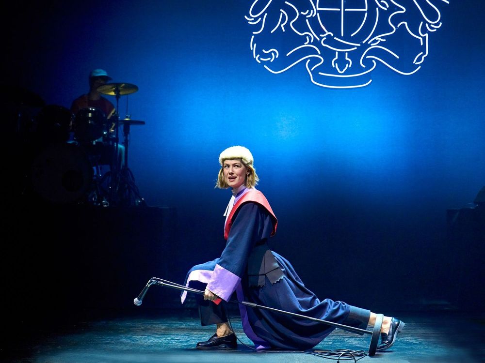 Rosamund Pike as Jessica, wearing a judge's wig and robes, dances with a microphone stand in front of the royal coat of arms. Inter Alia at the National Theatre's Lyttelton, 2025.