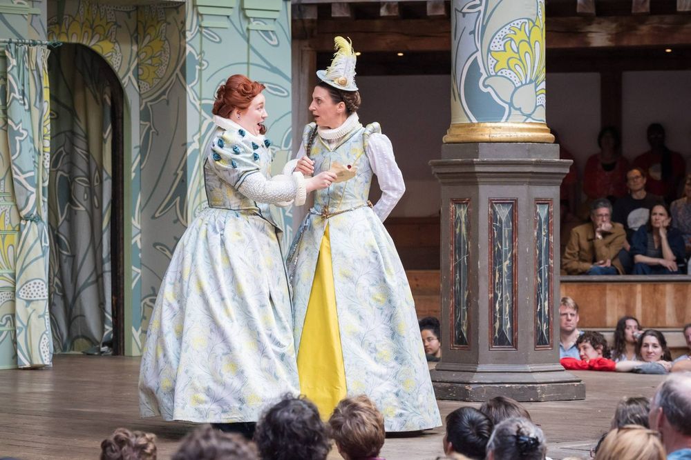 In white, patterned Elizabethan gowns, Katherine Pearce as Mistress Ford and Emma Pallant as Mistress Page chat animatedly. The Merry Wives of Windsor at Shakespeare's Globe, 2025.