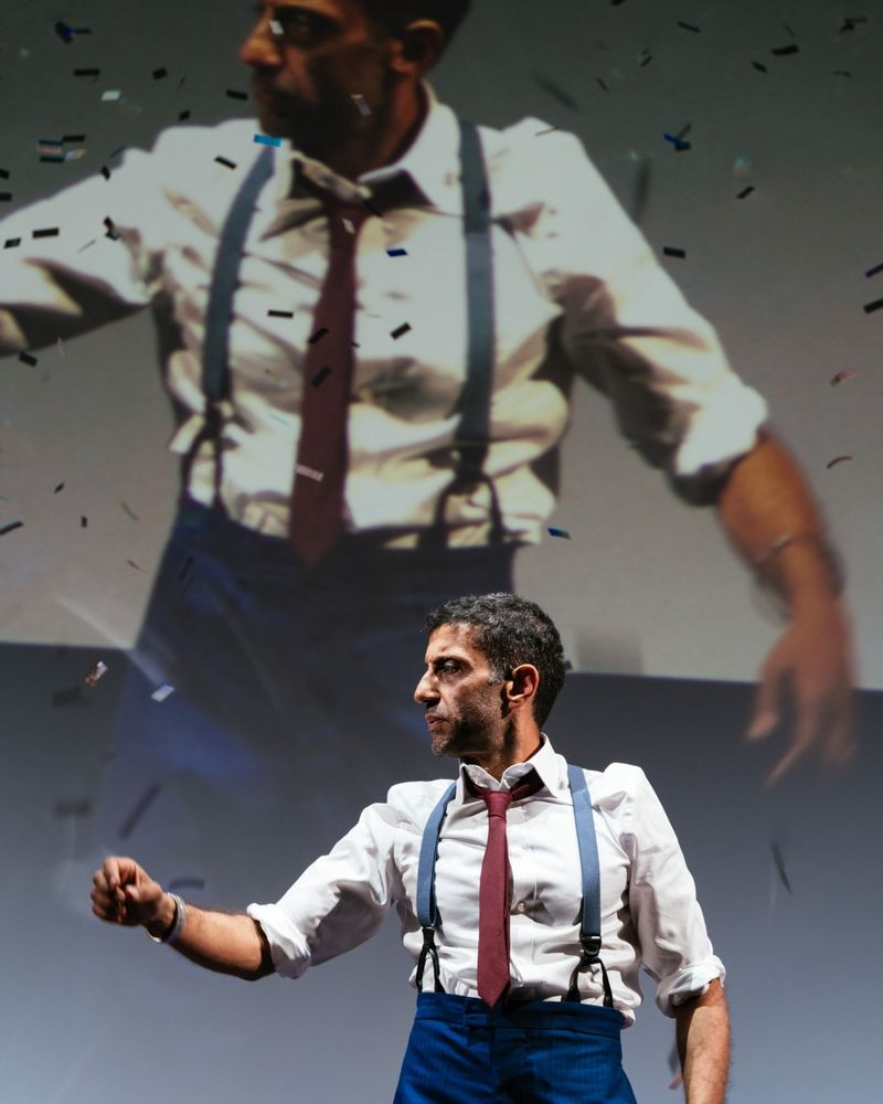Adeel Akhtar as Angad wears blue trousers and braces, a white shirt and red tie, standing in front of a huge scene showing him live, as at a political party conference. Confetti falls on him. The Estate at the National Theatre's Dorfman, 2025.