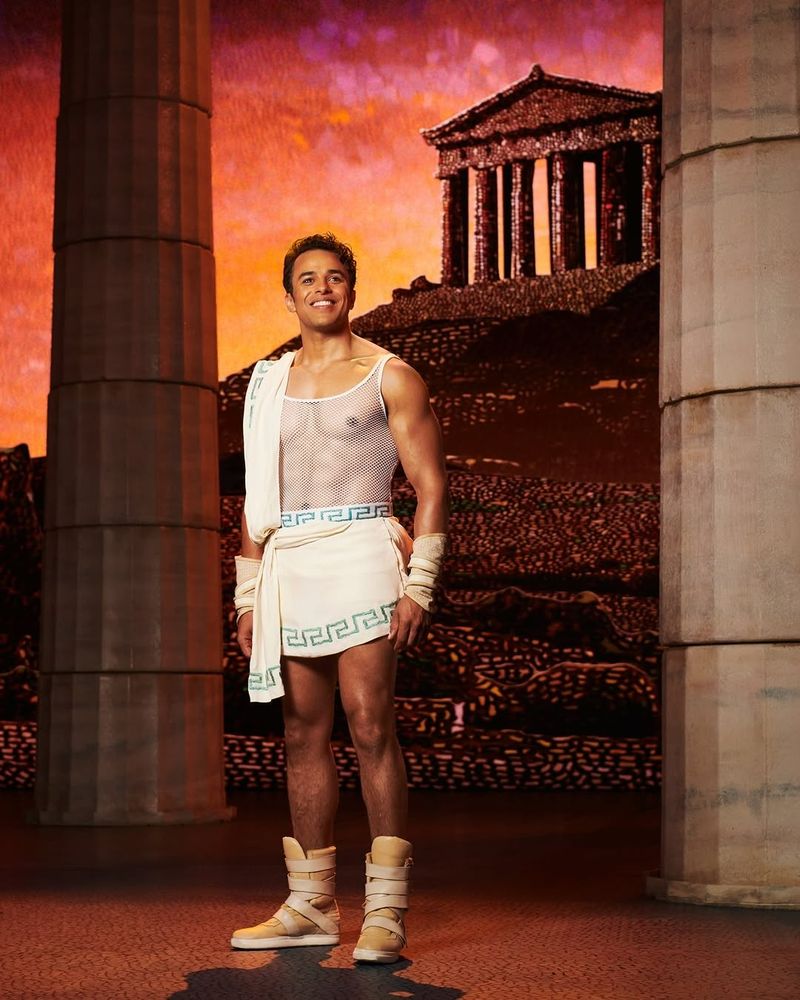 Luke Brady as Hercules in a tight mesh top. He's in front of a picture of the Acropolis, if anyone cares about that. Hercules at Theatre Royal Drury Lane, 2025.