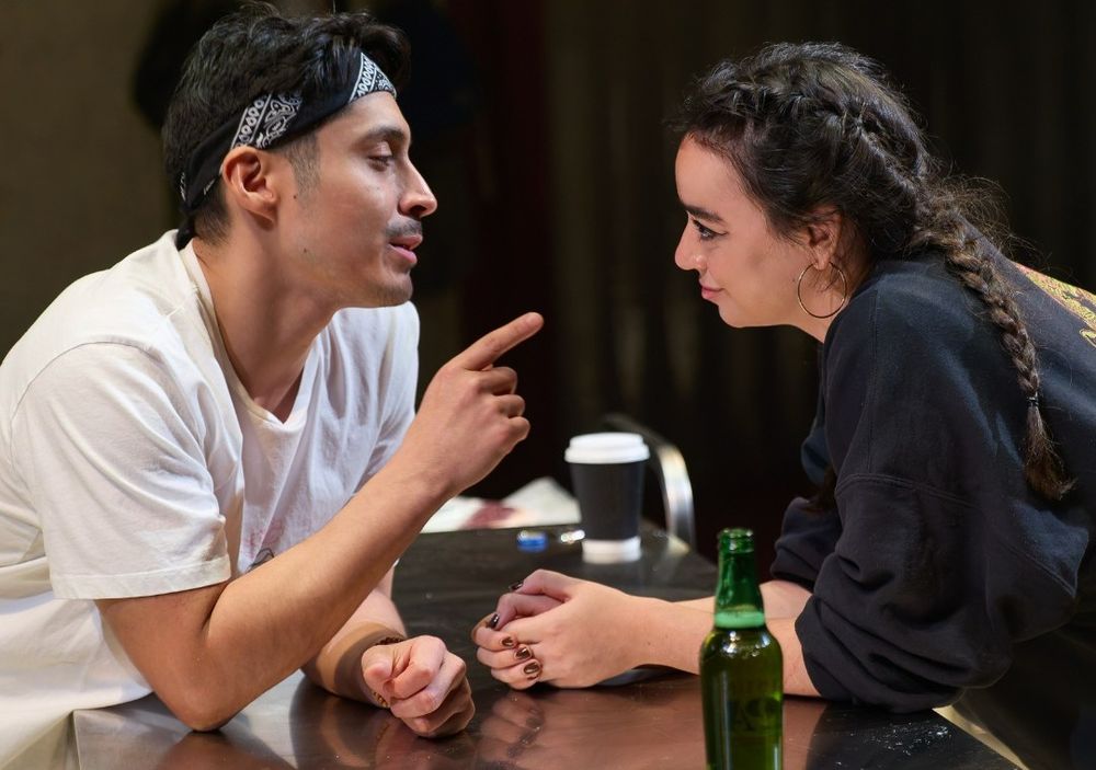 Marcello Cruz as JD and Mithra Malek as T lean across a counter, talking and gazing into each other's eyes. The Meat Kings Exclamation Mark Open Brackets Inc Full Stop Close Brackets of Brooklyn Heights at Park Theatre 200, 2025.