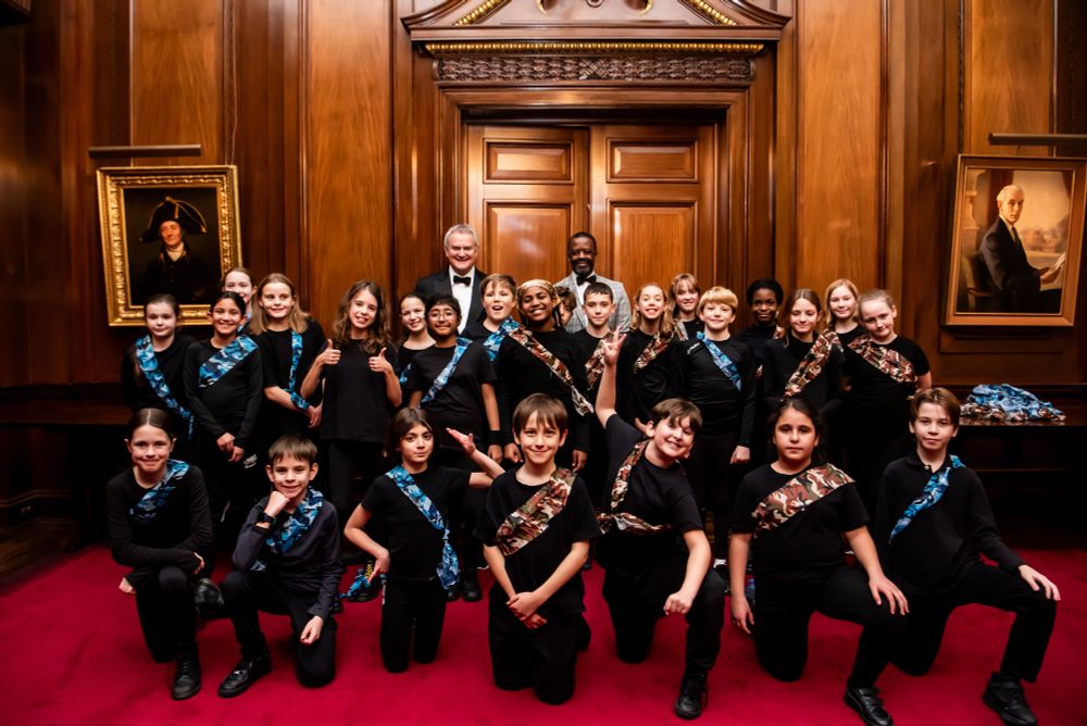 Photo of 25 children who are all in black and wearing either a blue or brown and green camouflage sash. Hugh Bonneville and Adrian Lester are standing behind them.