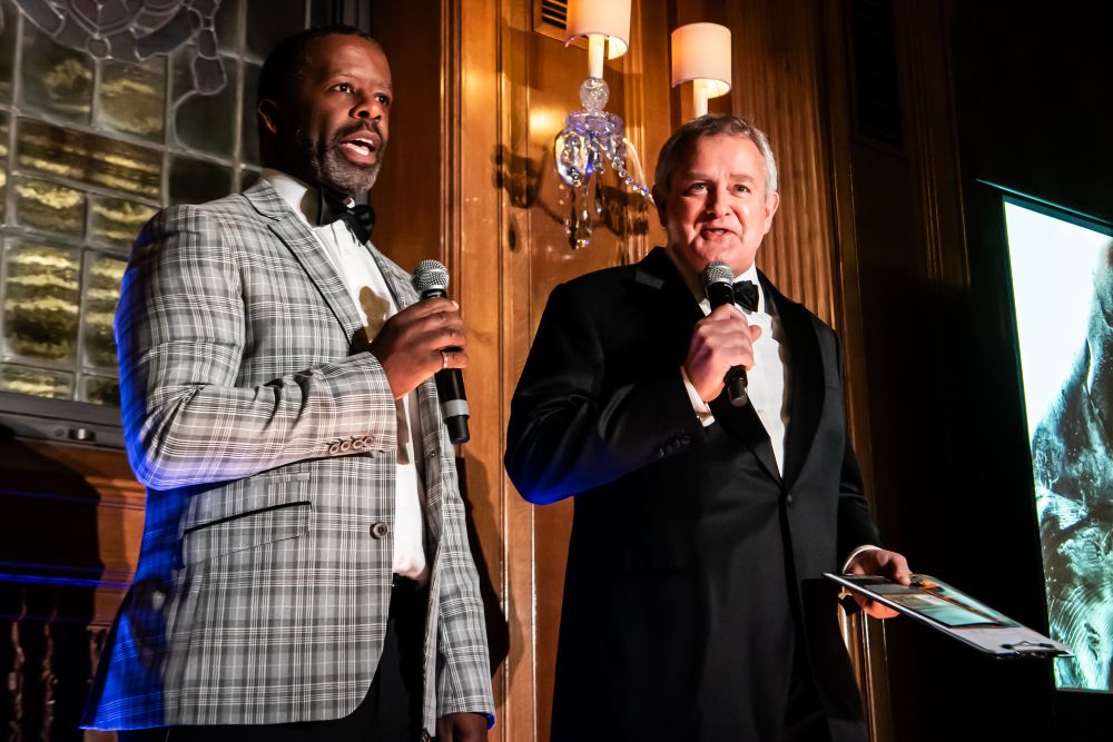Photo of actors Adrian Lester and Hugh Bonneville hosting the event. Both are holding microphones. Hugh is wearing a black tuxedo with a black bow tie and Adrian has on a grey check jacket with black trousers and a black bow tie.