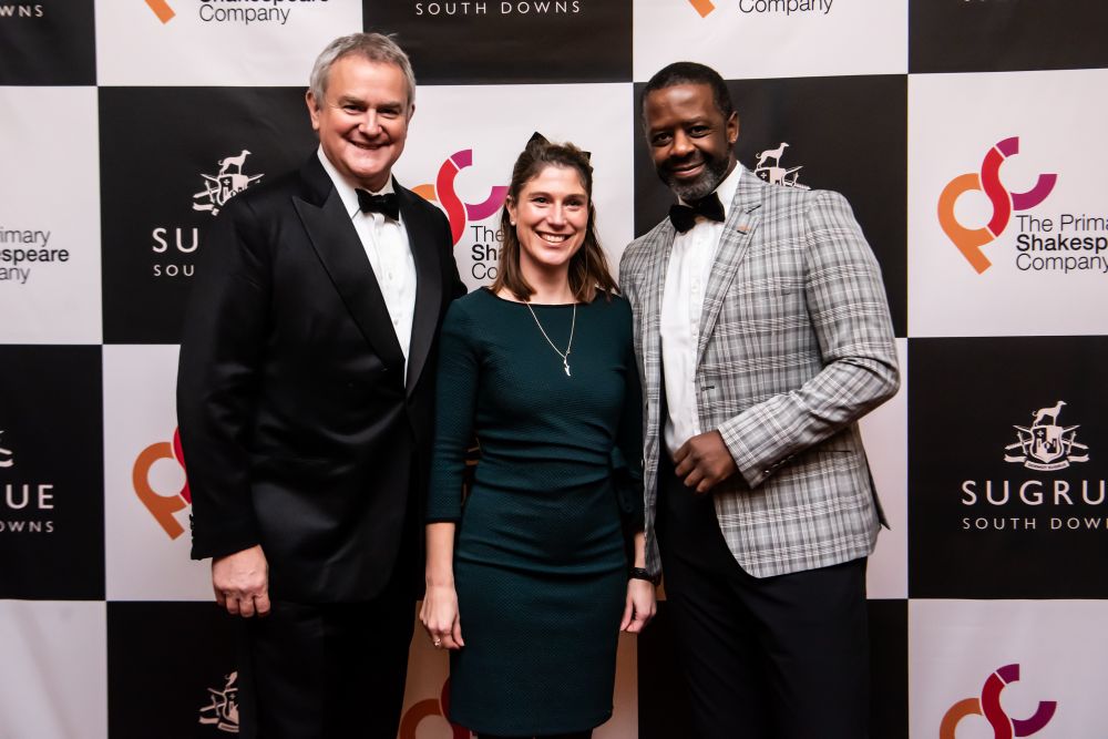 Photo of Hugh Bonneville, Ana Sugrue and Adrian Lester. Hugh is wearing a black tuxedo. Ana is wearing a dark green knee-length dress. Adrian is wearing a grey check jacket with black trousers and a black bow tie.