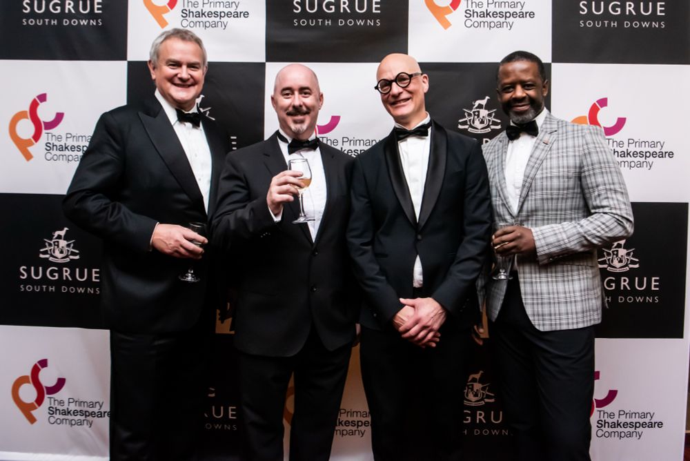 Photo of Hugh Bonneville, Neil Carter, Luke Hollowell-Williams and Adrian Lester. Hugh, Neil and Luke are wearing black tuxedos. Adrian is wearing a grey check jacket with black trousers and a black bow tie. Hugh, Neil and Adrian are holding glasses of wine.