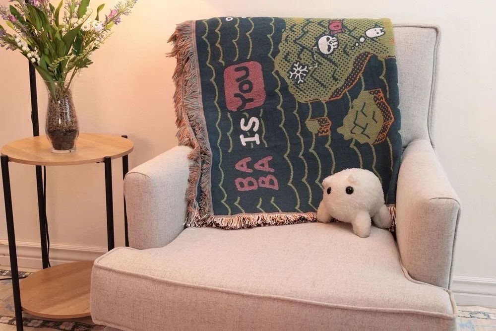 A blanket with the map from Baba Is You against a chair, with a Baba plushie sitting on the chair
