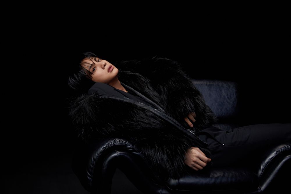 Jin of BTS is lying on a black leather chair. His hair is black and he’s wearing a black fur coat. His lips are even more beautiful than usual.