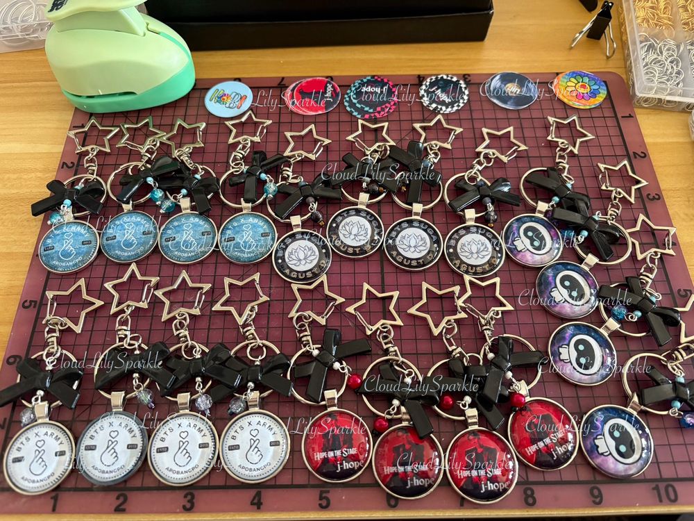 Handmade BTS inspired keychains designed by CloudLily. 5 finished designs laid out on a desk. Each keychain featuring black acrylic bows, star shaped spring hooks, keyring for keys, coordinating soarkly glass beads, and BTS inspired designs printed and set in metal bezel trays secured under glass domes. Soon to be available at CloudLilySparkle.Etsy.com