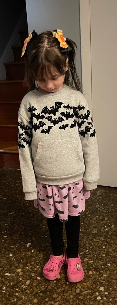 Young girl wear a gray heather sweatshirt with black felt bats over top a light purple cotton dress, also with black bats. Black stockings and bright pink crocs. Long brown hair is held back by two candy corn barrettes.