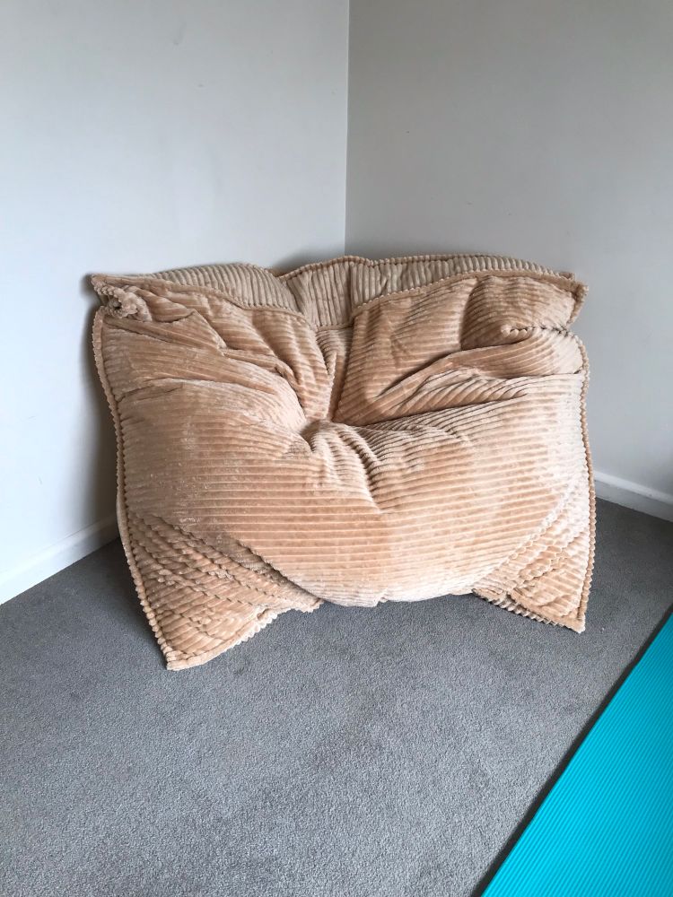 A large fawn corduroy beanbag in the corner of a room with pale walls, grey carpet. A strip of an aqua exercise mat appears on the bottom right of the image.
