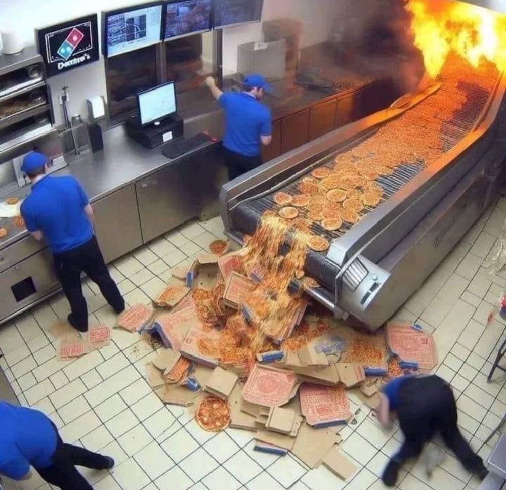 A complete catastrophe inside a Domino’s Pizza Kitchen with pizzas spilt all over the floor in a heap and someone presumably crying on the floor while everyone continues working 