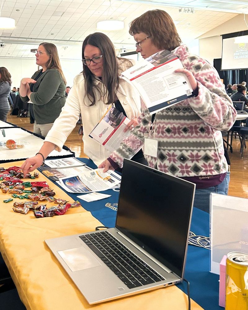 Two individuals are examining materials on a table at a conference, with various pamphlets and candies laid out in front of a laptop.