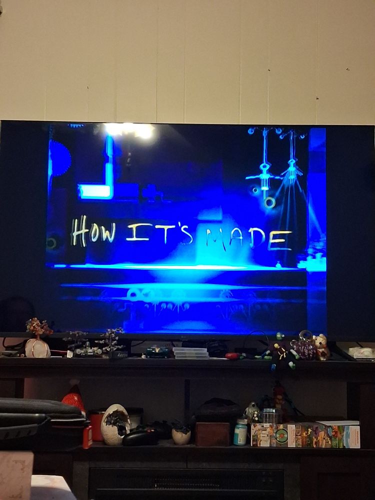 A picture of my television. You can see the title screen for the show How It's Made. There are knicknacks on the entertainment stand. The picture is taken from the angle of sitting on the couch looking at the tv.