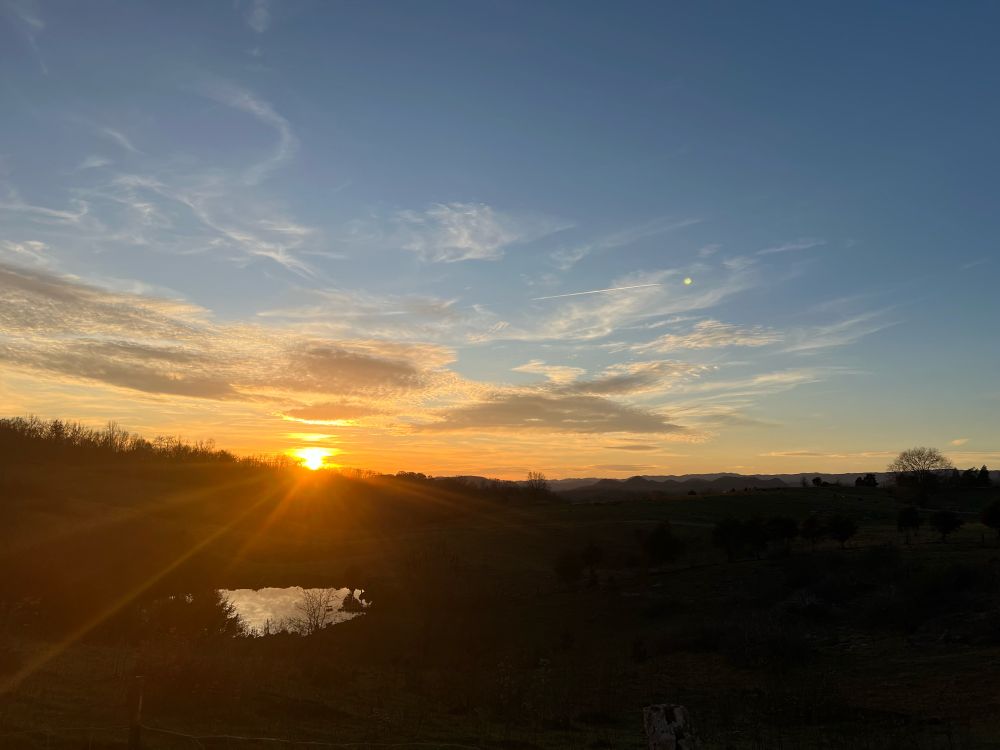 Sunset with mostly orange and yellow colors, blue sky at top. Silhouettes of mountains and some trees in bottom half of pic. Small pond on the left side. 