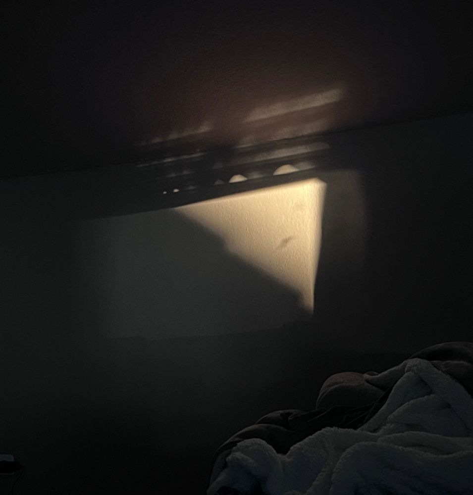 Eine Wand auf die das Licht des Fensters fällt. Man sieht ein Schatten von einem Dach und einen Fleck der präsent umgeben ist von Licht.