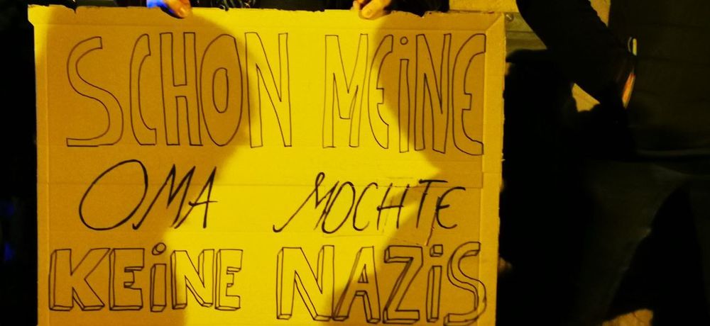 Zu sehen ist ein großes, aus Karton gebasteltes Schild, das jemand vor sich hält. Darauf steht in handgeschriebenen großen Buchstaben:
„SCHON MEINE OMA MOCHTE KEINE NAZIS“.
Die Person selbst ist kaum sichtbar, nur Hände und etwas Kleidung am Rand. Das Foto ist im Dunkeln aufgenommen, das Schild wird von gelblichem Licht beleuchtet.