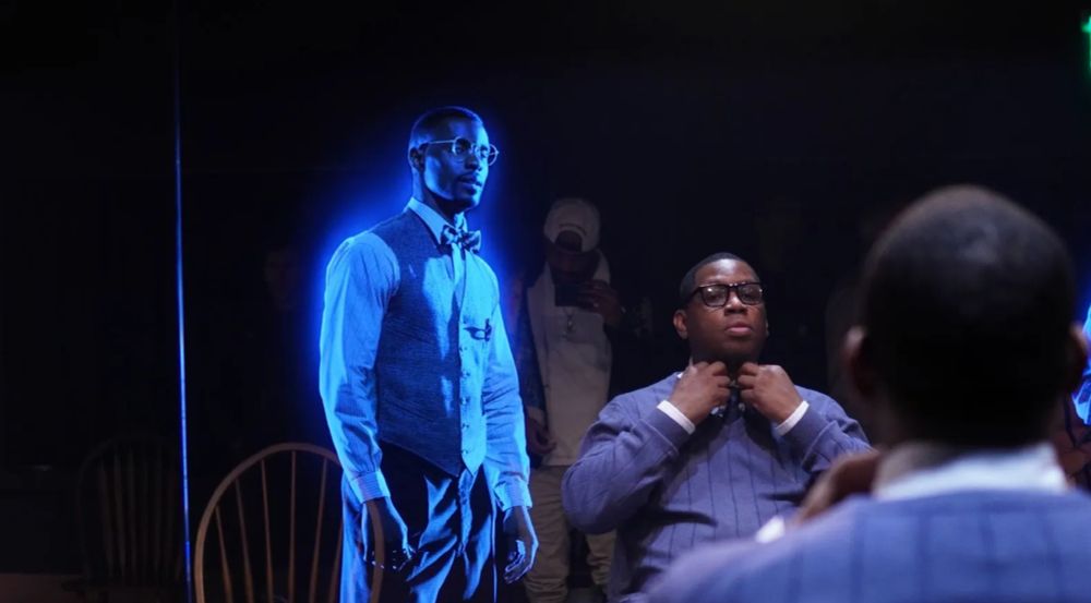 A man sits in a chair adjusting his bowtie while a digital barber hovers nearby. The photographer can be seen in the mirrored background.
