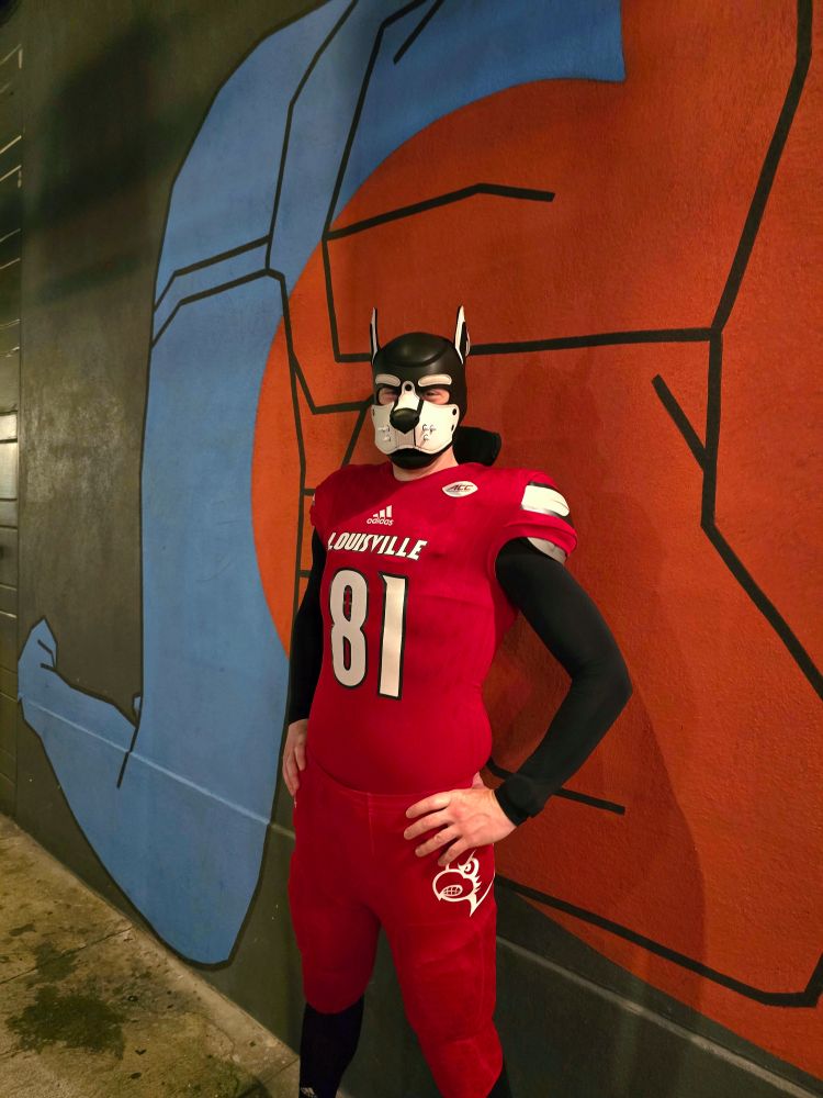 Loadz in pup white pup hood with spikes wearing red collegiate football gear and standing in front of a mural with hands on his hips and bent elbows looking at the camera. 