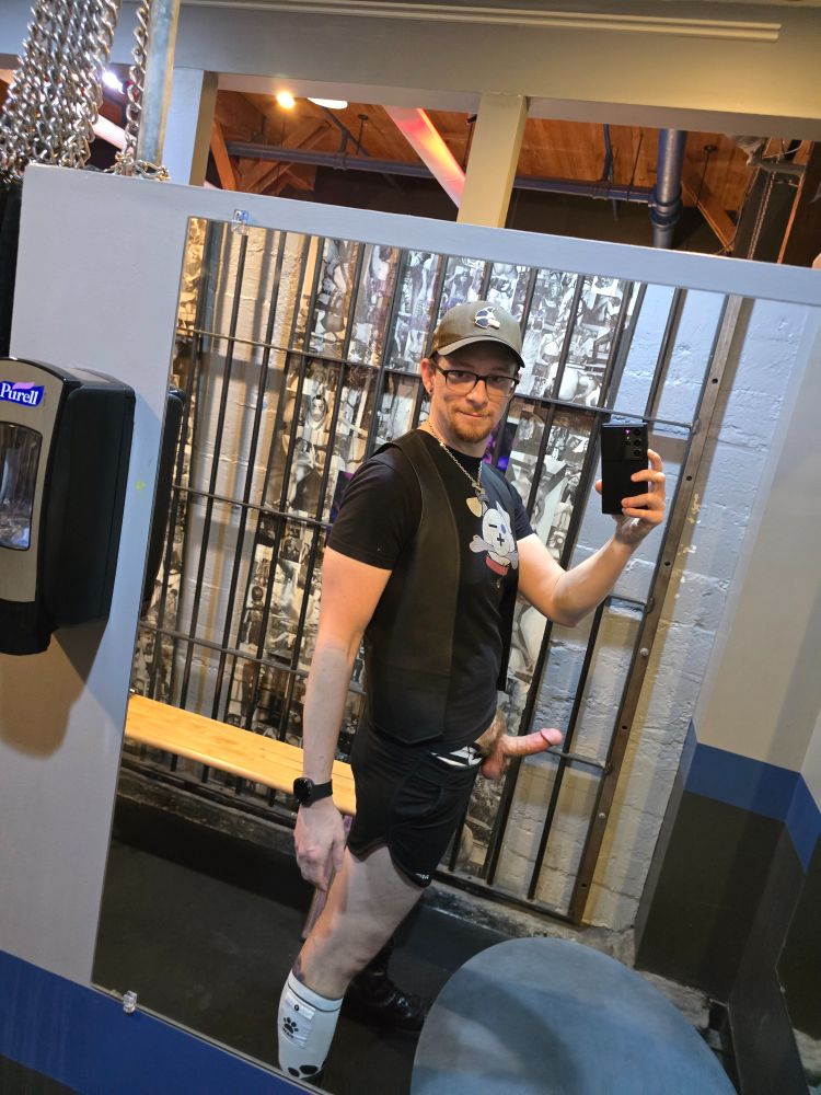 Loadz taking a selfie in the Mr S Leather dressing room with a hard cock while wearing a bar vest, black t-shirt, black MrS boots shorts, and pup hat.