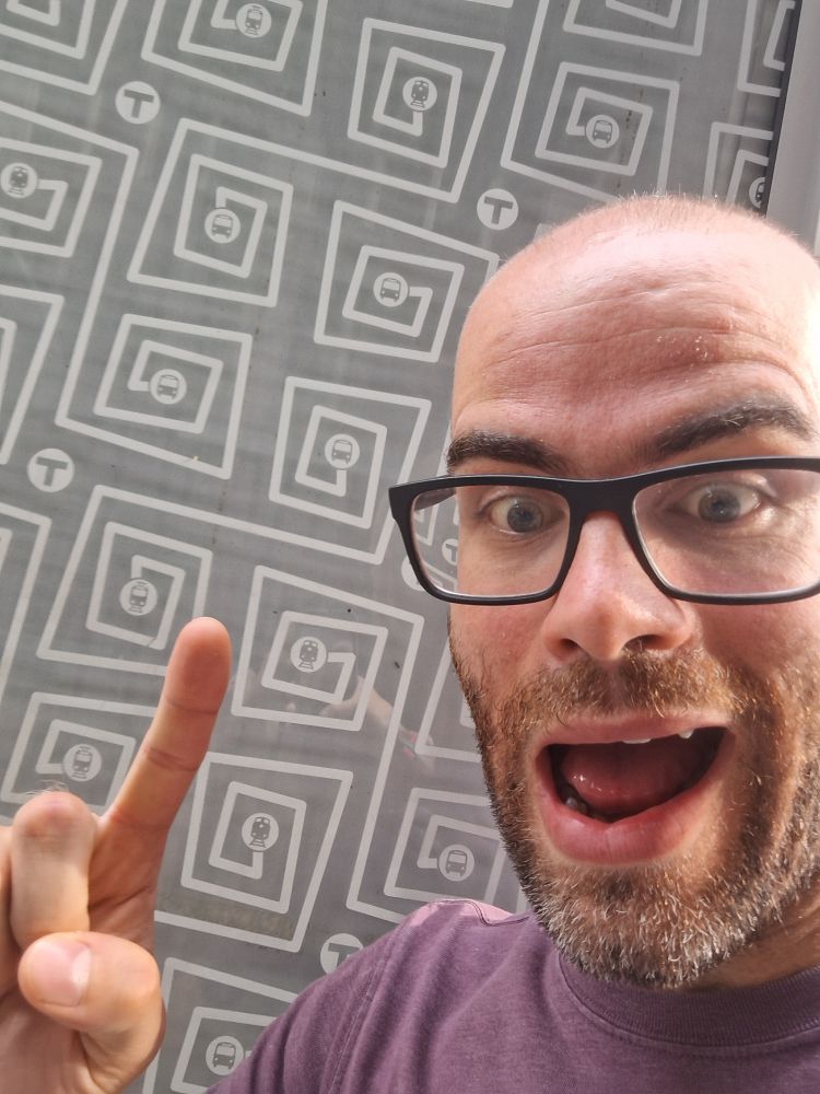 Photo of me using my finger to indicate the square spiral pattern of a glass bus shelter. My face is showing excitement, I guess?