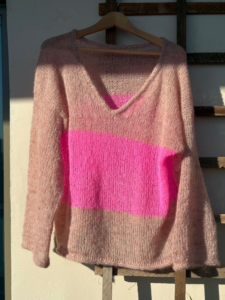 A handknit sweater hangs on a wooden hanger in the blazing sun. The sweater has a v-neck, long sleeves, and is mostly a dusty pink save for a thick bright hot pink stripe across the chest. 