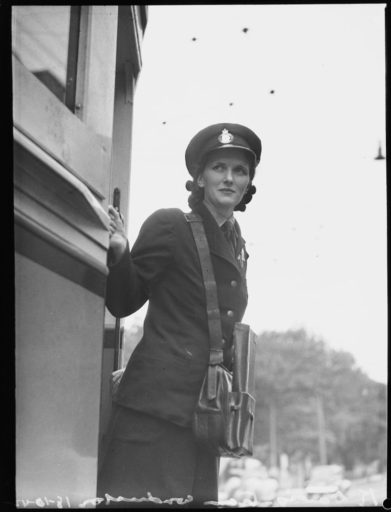 A behatted, uniformed woman with fare bag and ticket case, leans out of the door of a partially visible tram car. As the guard, she has a vigilant look