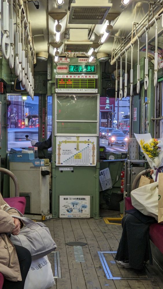 Looking forward in a tram. There are wooden floors; a green fares board obscuring the driver apart from his left hand, visible manipulating the control handle; two partially visible people dressed smartly with shopping and one with flowers, sit on opposite sides of the vehicle. Higher up are lights on the ceiling and long hanging straps