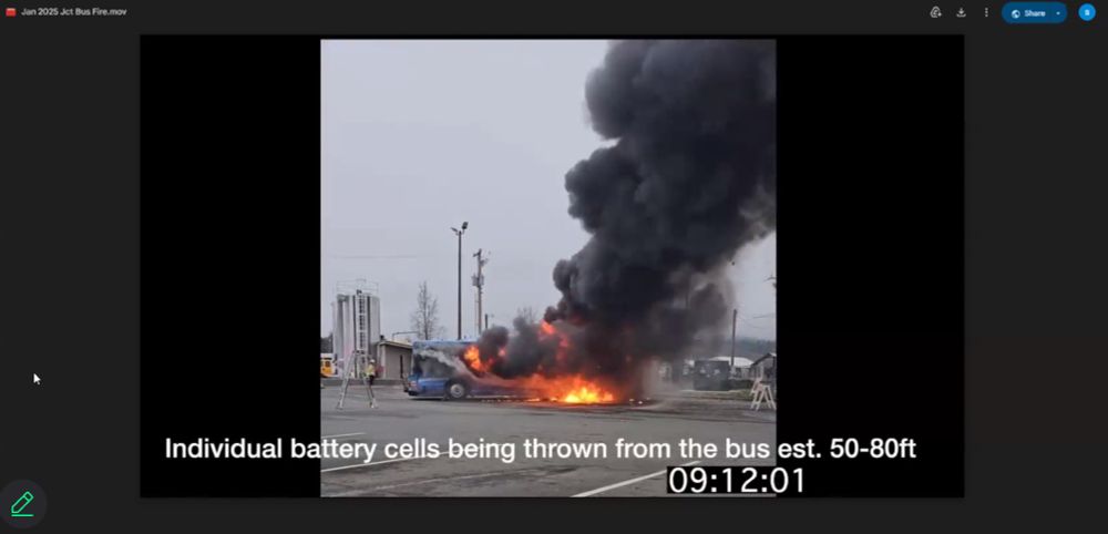 A screenshot of a blue electric bus on fire in a parking lot. There is a big black plume of smoke with gray skies behind. 