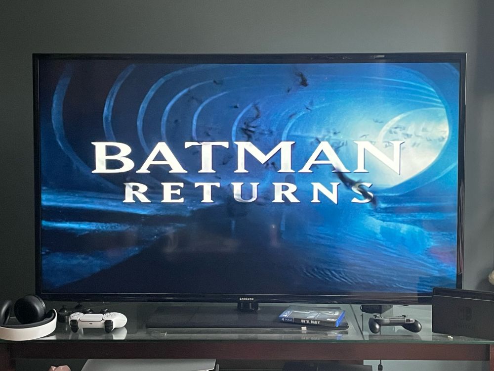 The title screen for Batman Returns, which features the text for the title and a background and a blue-lit sewer tunnel and bats flying toward the screen