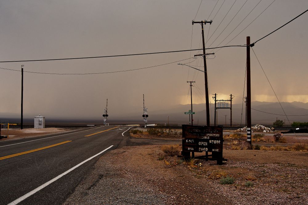 A two lane desert road near a small railroad town. The road starts on the left corner and goes away and towards the right to a railroad crossing with two barriers in the up position. The railroad is elevated so hides the road beyond. There are 4 wood power poles on the right side of the road and one on the right with wires running between them. On the right side of the road is an old wood sign, most of the white writing can't be made out. The parts that can be read are STORE OPEN BEER FOOD WINE beyond the wood sign is a street sign that says NIPTON DESERT RD. The horizon is storm clouds with a break in them on the right where the orange sky of sunset can be seen between mountains and the downpours of rain. On the left the mountains can't be seen and it looks like clouds from the horizon to the sky.