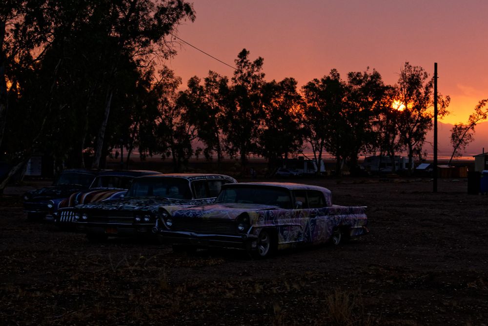 Four classic cars parked in the desert near some trees. The paint jobs on the cars are eccentric and not easy to describe. They are very colorful and not solid colors. The tops of the cars are shiny and reflecting the orange of the sunset through the clouds. Through the trees on the right of the frame the sun can be seen and it is fuzzy and orange. The whole sky is shades of orange.