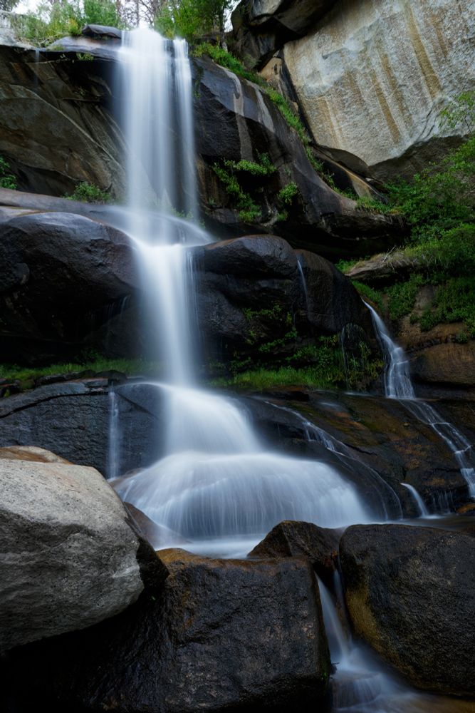 A waterfall blurred by a long exposure so that it is a dreamy feathery white falling from the top of grey black rocky wall of a valley. The waterfall is broken in multiple places from near the top of the frame to the bottom. There are what appears to be pine trees at the top of the waterfall and on the right of the frame. There is green vegetation dispersed through the rocks as well as moss.