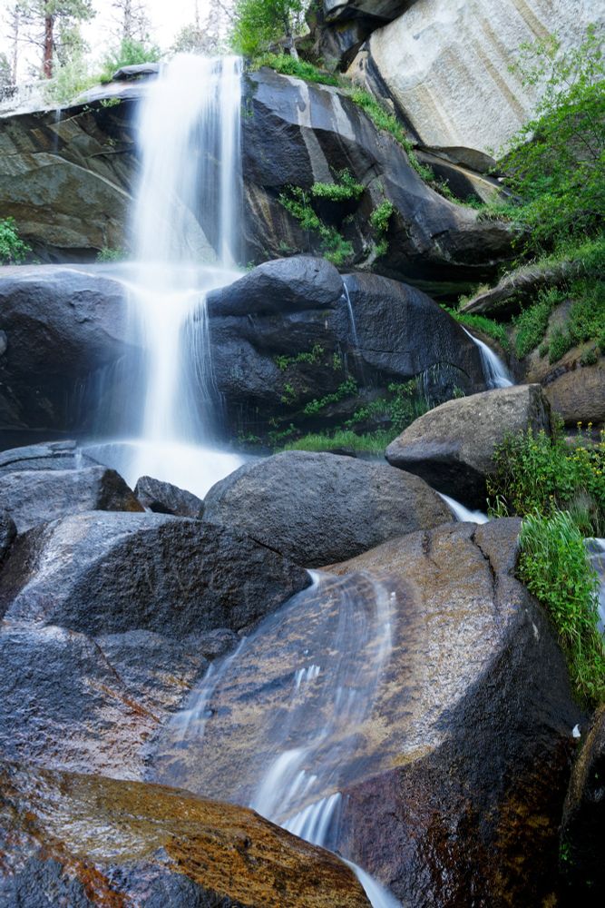 A waterfall blurred by a long exposure so that it is a dreamy feathery white falling from the top of grey black rocky wall of a valley. The waterfall is broken in multiple places from near the top of the frame to the bottom. There are what appears to be pine trees at the top of the waterfall. There is green vegetation dispersed through the rocks as well as moss. The rocks are various colors and shades from a nearly white where they are completely dry on the top right and left. Where the rocks are wet they range in color from rust on the foreground rocks  which lead to a tan moving up the frame to dark grey and a coal color at the middle of the frame and directly around the start of the waterfall at the top of the frame.