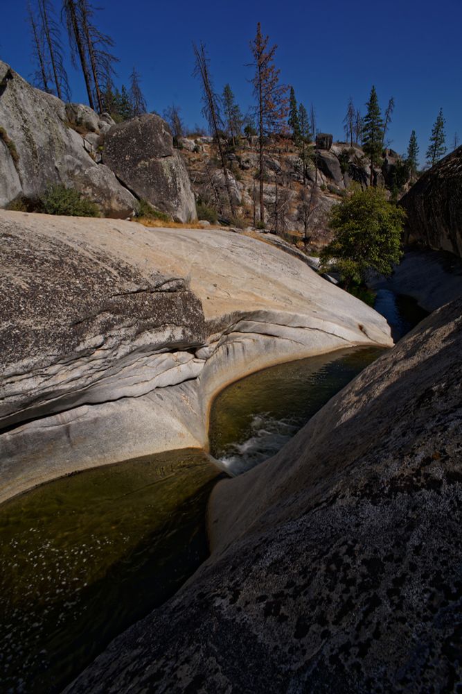 A picture from above and to the side of a creek that carved out pools in a rock outcrop. The outcrop is a mix of whites, greys, and tans. There are two visible oval pools with a small waterfall between them, after the second pool water can be seen in the distance at a lower level. The closer part of the outcrop has a steep drop and is mostly in shadow. roughly two thirds of the way across the pools and on the other side of them it is sunny. There is vegetation that is gray and brown on the far side of the creek growing past the rocky outcrop and in the distance. The sky above is a deep blue and takes up about 1/6th of the frame at the top.