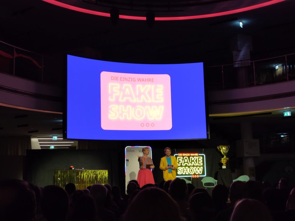 Showbühne mit einer Moderatorin im bunten Kleid und einem Moderator im gelben Anzug. Im Vordergrund ist das Publikum von hinten zu sehen. Der Bildschirm zeigt die Aufschrift: Die einzig wahre Fakeshow 
