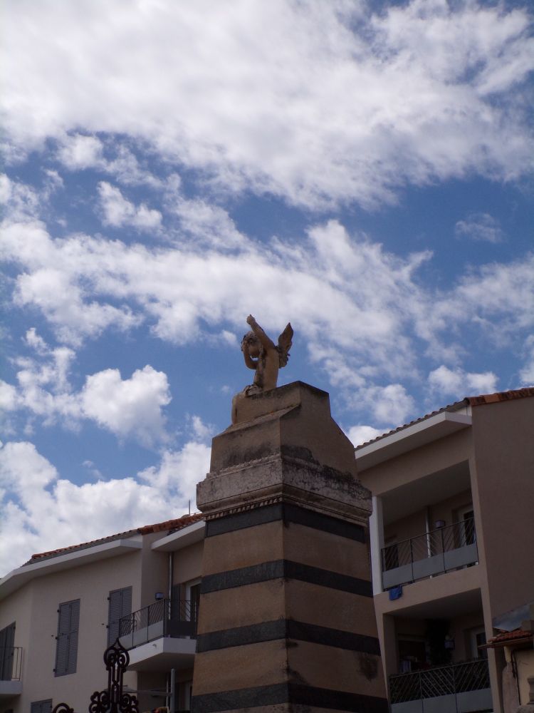 Une petite statue de chérubin qui surmonte une possible sépulture, vue d'en bas et de trois quart. Il détonne avec en fond le ciel bleu parsemé de nuage, donnant l'impression d'en faire partie.